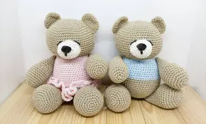 Due orsetti teddy amigurumi, uno con vestitino rosa e l'altro con maglioncino azzurro, seduti su un tavolino di legno.