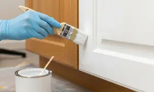 Mano con guanto blu dipinge una porta bianca con un pennello. Vicino, una lattina di vernice bianca. Impara come diluire la pittura per un risultato perfetto.