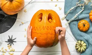 Mani che scolpiscono una zucca per Halloween, creando un volto spaventoso. Un gatto nero e un teschio completano l'atmosfera.