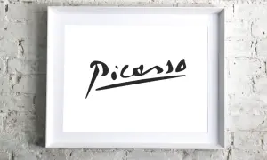 Firma pittore su tela, un'iconica firma di Picasso in nero su sfondo bianco, incorniciata in bianco, appesa a un muro di mattoni bianchi.