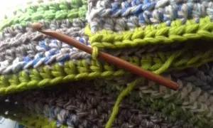 Coperta ai ferri con avanzi di lana, un'opera in corso con un uncinetto di legno che lavora maglie verdi su un tessuto multicolore.