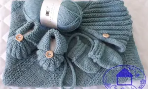 Un set per neonato ai ferri: copertina, scarpine e cappellino in lana merino azzurra, con bottoncini in legno.