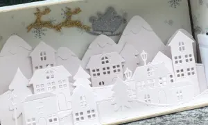 Sagome di un villaggio di Natale da ritagliare, con renne e slitta di Babbo Natale che volano sopra le casette innevate.