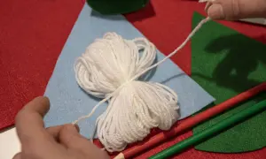 Mani che preparano lavoretti di Natale con la lana facili: gomitolo bianco, feltro colorato e matite.
