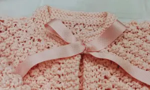 Dettaglio di un golfino bimba ai ferri, rosa pastello, con fiocco in raso. Spiegazioni per realizzarlo.