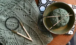 Lavori con lana sfumata: un maglione traforato, ferri da maglia, un gomitolo in una ciotola e un quaderno con motivo esagonale.