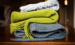 Pile di sciarpe ai ferri: una grigia, una verde con frange e una grigio chiaro in cima. Quanti punti per una sciarpa ai ferri? Dipende dalla lana e dalla maglia!