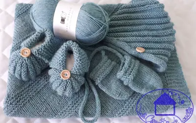 Un set per neonato ai ferri: copertina, scarpine e cappellino in lana merino azzurra, con bottoncini in legno.
