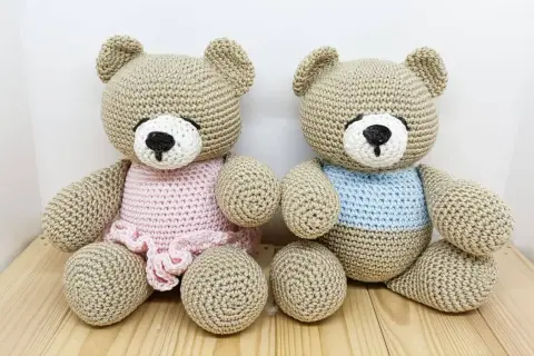 Due orsetti teddy amigurumi, uno con vestitino rosa e l'altro con maglioncino azzurro, seduti su un tavolino di legno.