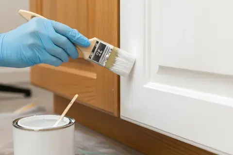 Mano con guanto blu dipinge una porta bianca con un pennello. Vicino, una lattina di vernice bianca. Impara come diluire la pittura per un risultato perfetto.