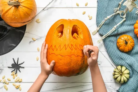 Mani che scolpiscono una zucca per Halloween, creando un volto spaventoso. Un gatto nero e un teschio completano l'atmosfera.