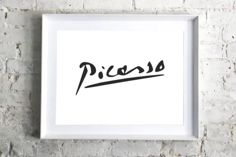 Firma pittore su tela, un'iconica firma di Picasso in nero su sfondo bianco, incorniciata in bianco, appesa a un muro di mattoni bianchi.