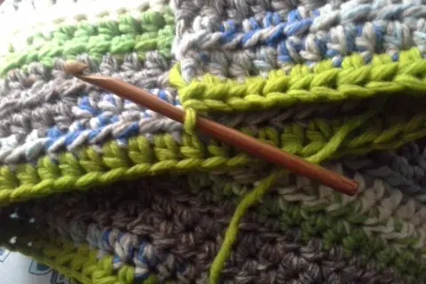 Coperta ai ferri con avanzi di lana, un'opera in corso con un uncinetto di legno che lavora maglie verdi su un tessuto multicolore.