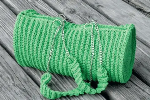 Borsa verde brillante lavorata all'uncinetto doppia punta, con manici e catene argentate, adagiata su assi di legno.