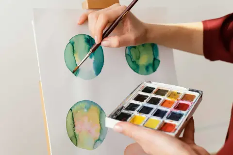 Mano che dipinge cerchi verdi e gialli con acquerello su tela. Palette di colori in primo piano.