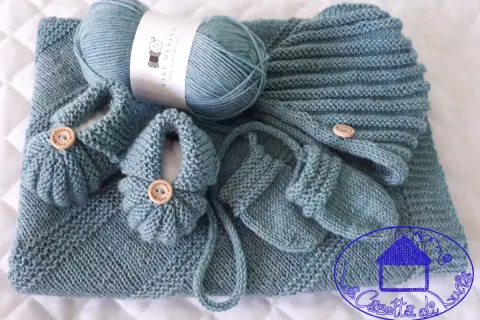 Un set per neonato ai ferri: copertina, scarpine e cappellino in lana merino azzurra, con bottoncini in legno.