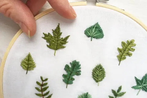 Mani che tengono un telaio da ricamo, con diverse piccole foglie ricamate su tessuto bianco. Un'arte delicata per ricamare foglie piccole.