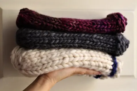 Tre berretti in lana ai ferri, uno bordeaux, uno grigio e uno bianco con striscia blu, impilati su una mano.