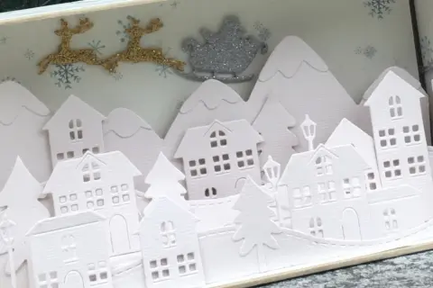 Sagome di un villaggio di Natale da ritagliare, con renne e slitta di Babbo Natale che volano sopra le casette innevate.