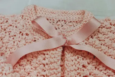 Dettaglio di un golfino bimba ai ferri, rosa pastello, con fiocco in raso. Spiegazioni per realizzarlo.