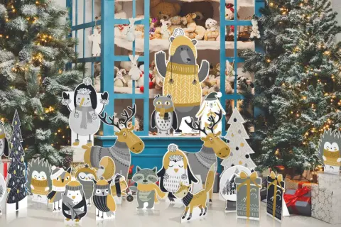 Sagome natalizie grandi di animali e alberi, con un orso al centro, davanti a un albero di Natale innevato e una vetrina piena di peluche.