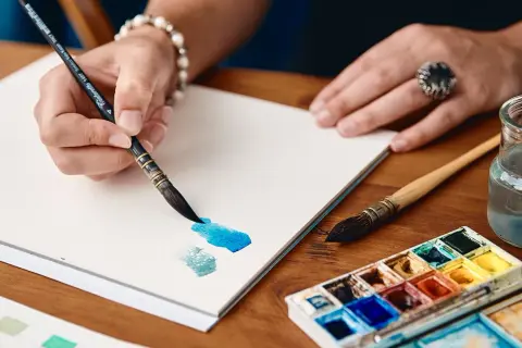 Mano che dipinge con acquerelli su un sottilissimo foglio per acquerelli e litografie. Tavolozza di colori e pennelli vicini.