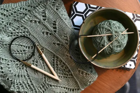 Lavori con lana sfumata: un maglione traforato, ferri da maglia, un gomitolo in una ciotola e un quaderno con motivo esagonale.