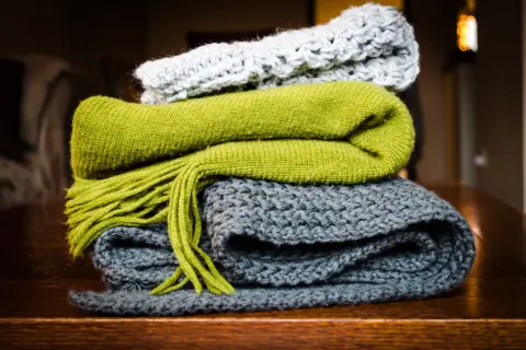 Pile di sciarpe ai ferri: una grigia, una verde con frange e una grigio chiaro in cima. Quanti punti per una sciarpa ai ferri? Dipende dalla lana e dalla maglia!