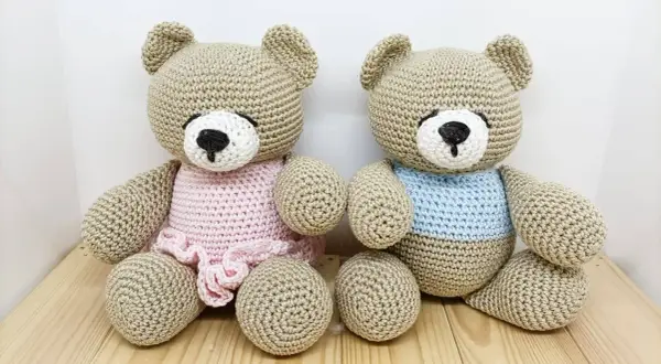 Due orsetti teddy amigurumi, uno con vestitino rosa e l'altro con maglioncino azzurro, seduti su un tavolino di legno.