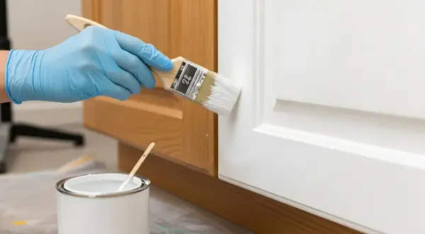 Mano con guanto blu dipinge una porta bianca con un pennello. Vicino, una lattina di vernice bianca. Impara come diluire la pittura per un risultato perfetto.