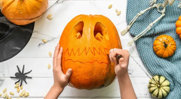 Mani che scolpiscono una zucca per Halloween, creando un volto spaventoso. Un gatto nero e un teschio completano l'atmosfera.