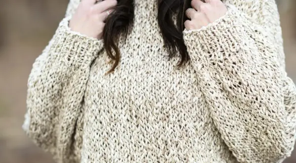 Primo piano di un maglione a maglia color crema con filato a trama aperta. Le mani della donna, adornate con anelli, giocano con i suoi capelli scuri.