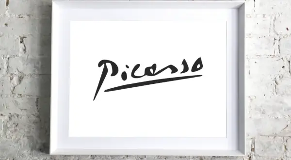 Firma pittore su tela, un'iconica firma di Picasso in nero su sfondo bianco, incorniciata in bianco, appesa a un muro di mattoni bianchi.