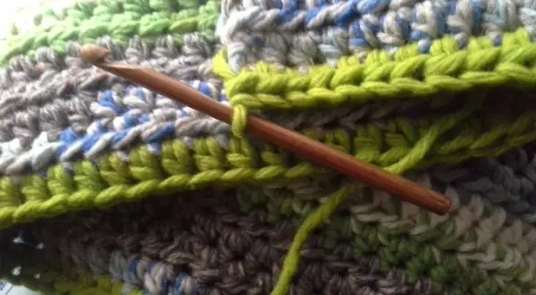 Coperta ai ferri con avanzi di lana, un'opera in corso con un uncinetto di legno che lavora maglie verdi su un tessuto multicolore.