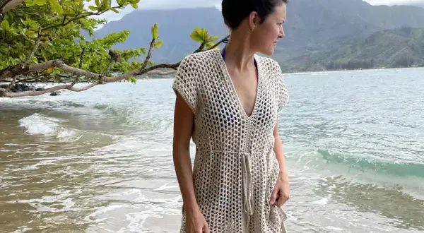 Donna in copricostume uncinetto beige, con schema gratis, sulla spiaggia con montagne sullo sfondo.