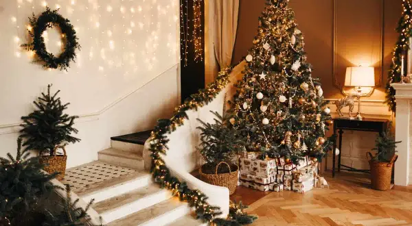 Un albero di Natale scintillante, decorato con nastri natalizi per albero, addobbi dorati e luci calde. La scala è adornata da ghirlande luminose e piccoli abeti in cesti.