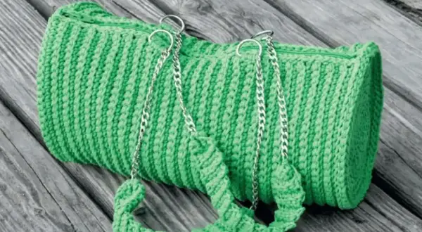 Borsa verde brillante lavorata all'uncinetto doppia punta, con manici e catene argentate, adagiata su assi di legno.