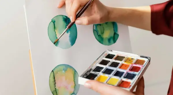 Mano che dipinge cerchi verdi e gialli con acquerello su tela. Palette di colori in primo piano.
