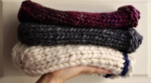 Tre berretti in lana ai ferri, uno bordeaux, uno grigio e uno bianco con striscia blu, impilati su una mano.