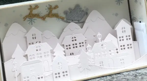 Sagome di un villaggio di Natale da ritagliare, con renne e slitta di Babbo Natale che volano sopra le casette innevate.