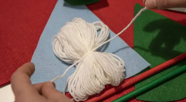 Mani che preparano lavoretti di Natale con la lana facili: gomitolo bianco, feltro colorato e matite.