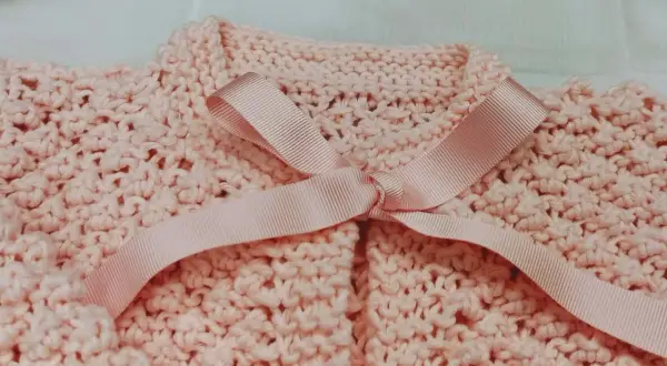 Dettaglio di un golfino bimba ai ferri, rosa pastello, con fiocco in raso. Spiegazioni per realizzarlo.