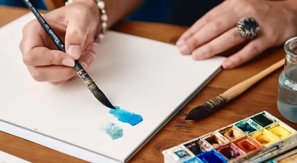 Mano che dipinge con acquerelli su un sottilissimo foglio per acquerelli e litografie. Tavolozza di colori e pennelli vicini.