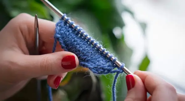 Mani con unghie rosse lavorano a maglia un filato blu. Impara come lavorare la lana con questo dettaglio ravvicinato.