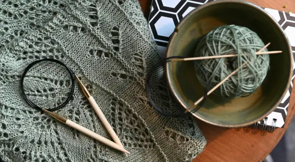 Lavori con lana sfumata: un maglione traforato, ferri da maglia, un gomitolo in una ciotola e un quaderno con motivo esagonale.