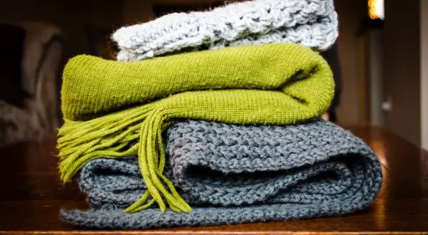 Pile di sciarpe ai ferri: una grigia, una verde con frange e una grigio chiaro in cima. Quanti punti per una sciarpa ai ferri? Dipende dalla lana e dalla maglia!
