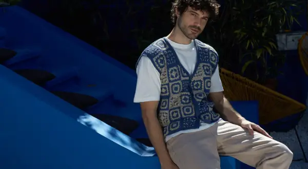 Uomo con riccio gilet senza maniche all'uncinetto blu e beige, seduto su una struttura blu.