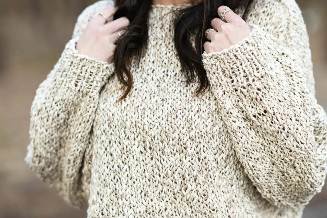 Primo piano di un maglione a maglia color crema con filato a trama aperta. Le mani della donna, adornate con anelli, giocano con i suoi capelli scuri.