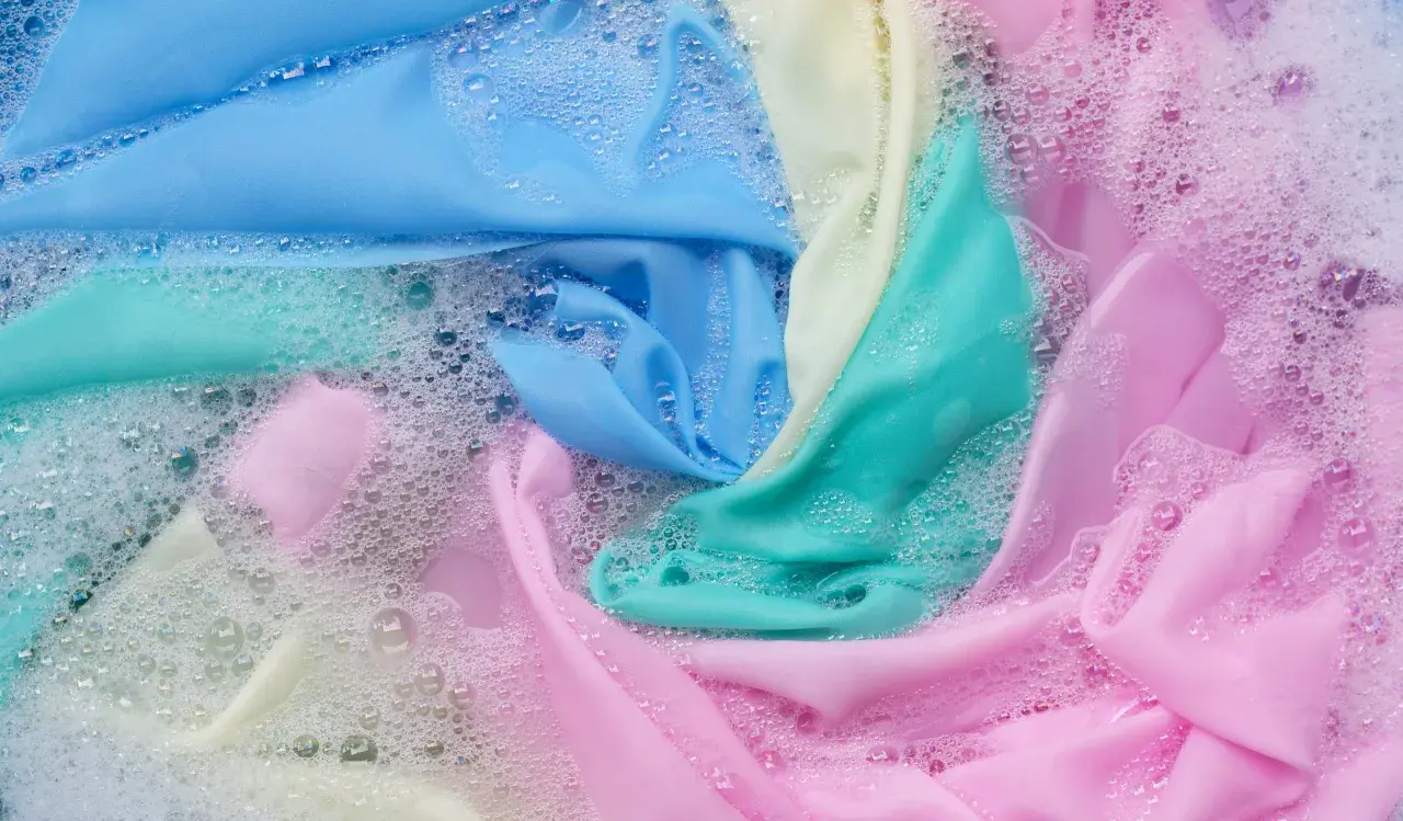 Tessuti color pastello, tra cui un **acetato tessuto** azzurro, si muovono tra bolle di sapone in un lavaggio delicato.