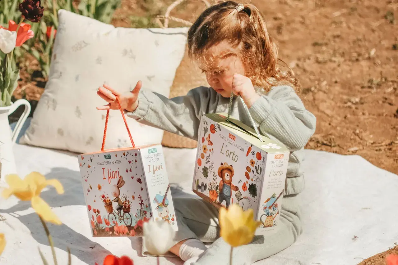Bambina con due scatole di puzzle ad acqua, perfetti per le attività estive. Un coniglietto in bici e un orsetto giardiniere animano i giochi.